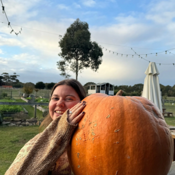 Smiling woman holding a giant pumpkin outdoors on a farm with garden beds and string lights.